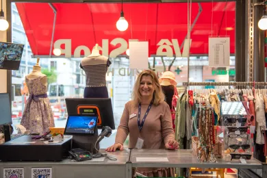 Une collaboratrice souriante se tient derrière la caisse d’une boutique de vêtements de seconde main de la Croix-Rouge genevoise, entourée de vêtements et d’accessoires.